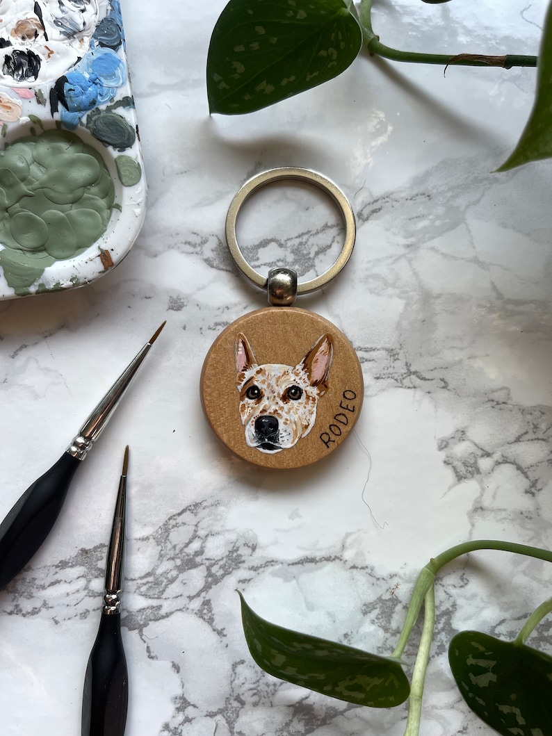 May include: A wooden keychain with a painted portrait of a dog. The dog is a brown and white Australian Cattle Dog with a black nose. The keychain is round and has a silver ring attached. The dog's name, "RODEO", is written in white on the keychain.