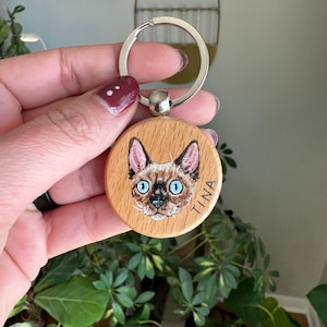 May include: A round wooden keychain with a painted portrait of a cat with blue eyes. The cat's name, "TINA", is written on the keychain.