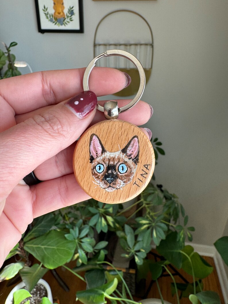 May include: A round wooden keychain with a painted portrait of a cat with blue eyes. The cat's name, "TINA", is written on the keychain.