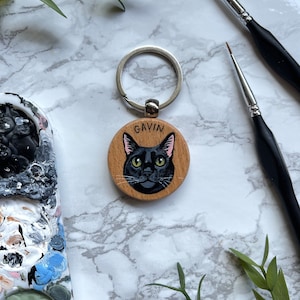 May include: A wooden keychain with a black cat painted on it. The cat has green eyes and a pink nose. The keychain is attached to a silver ring. The name "GAVIN" is written on the wood.