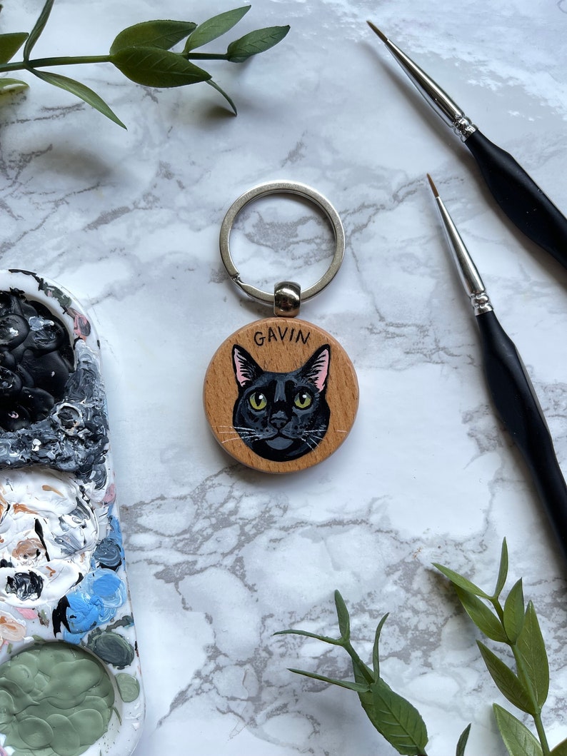 May include: A wooden keychain with a black cat painted on it. The cat has green eyes and a pink nose. The keychain is attached to a silver ring. The name "GAVIN" is written on the wood.