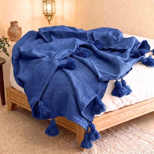 Peut inclure: Un jeté de lit bleu roi vibrant avec une surface en tissu texturé, orné de multiples pompons bleus assortis fixés le long des bords. La couverture est drapée sur un drap blanc, mettant en valeur son aspect doux et plissé.