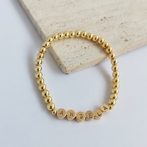 May include: A gold beaded bracelet with the name "ANGELA" spelled out in gold letters.
