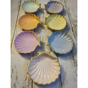 May include: Seven seashell-shaped dishes in pastel shades of pink, blue, yellow, and orange. Each dish has a gold trim. The dishes are arranged on a white wooden surface. The dishes are ideal for serving small portions.