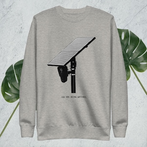 May include: A light gray sweatshirt featuring a black and white graphic of a surveillance device. The text "YOU ARE BEING WATCHED" is printed in black below the graphic. The sweatshirt is made of a soft material.