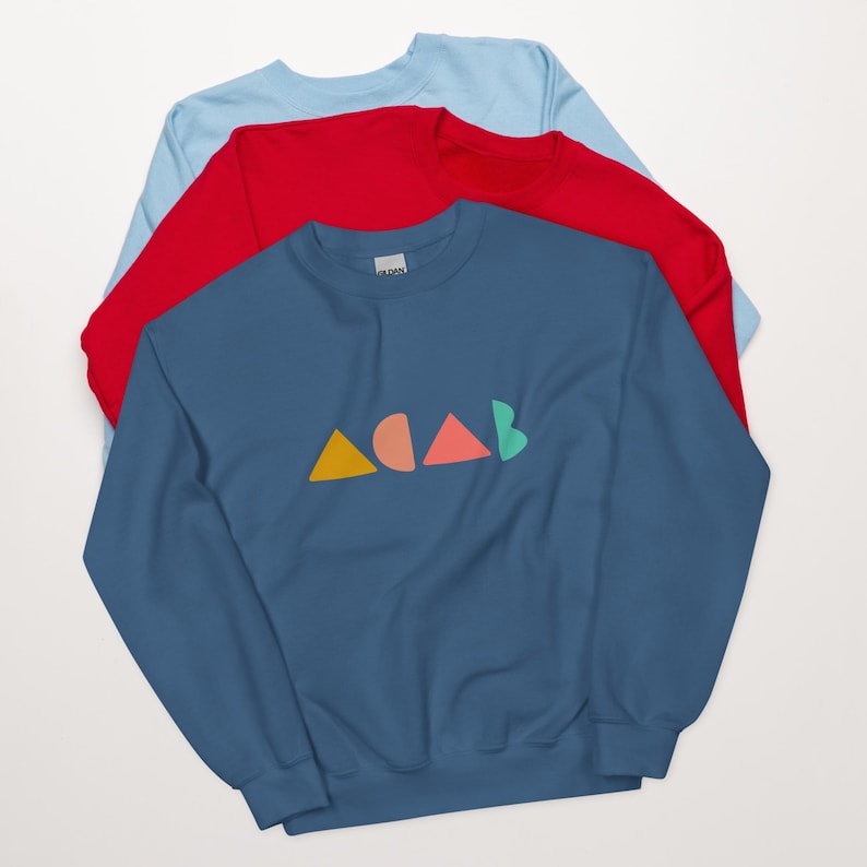 K&ouml;nnte beinhalten: Drei &uuml;bereinander gelegte Sweatshirts in Blau, Rot und Hellblau. Das vorderste blaue Sweatshirt zeigt den Text "ACAB" in farbenfrohen geometrischen Formen. Der Text besteht aus einem gelben Dreieck, einem pfirsichfarbenen Halbkreis, einem korallenfarbenen Dreieck und einem gr&uuml;nen Halbkreis.
