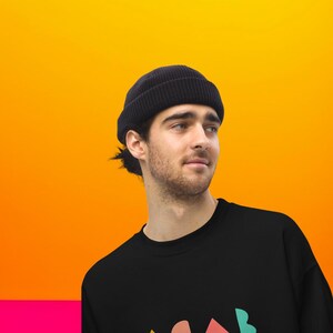 K&ouml;nnte beinhalten: Eine schwarze Beanie-M&uuml;tze und ein schwarzes Sweatshirt mit einem farbenfrohen, abstrakten Design. Die M&uuml;tze ist aus geripptem Strickmaterial. Der Hintergrund ist ein Farbverlauf aus Orange und Gelb.