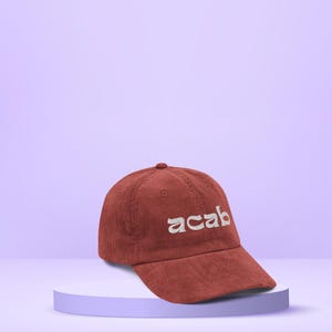 May include: A terracotta-coloured corduroy baseball cap with the word "acab" embroidered in a light cream colour. The cap is displayed on a light purple pedestal against a light purple background. The cap has a curved brim and a rounded crown.