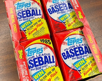 May include: A box of 1985 Topps Major League Baseball picture cards with bubble gum. The red and yellow packs are labeled with the Topps logo and the words "Baseball" and "Win". The box is open, revealing the packs.