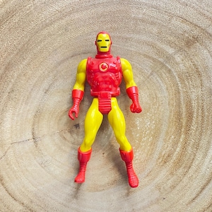 May include: A vintage Iron Man action figure. The figure is primarily yellow with red accents on the chest, boots, gloves, and helmet. The figure is standing on a wooden surface.