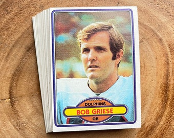 May include: A vintage football trading card featuring Bob Griese, quarterback for the Miami Dolphins. The card is white with blue, orange, and yellow accents. It features a photo of Griese in his Dolphins uniform and the text "Dolphins, Bob Griese, QB".