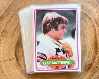 May include: A vintage football trading card featuring a photo of Clay Matthews, a linebacker for the Cleveland Browns. The card has a pink border and orange text that reads "Cleveland Browns" and "Clay Matthews LB".