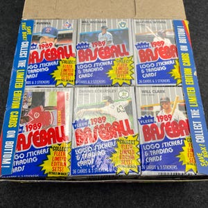 May include: A box of 1989 Fleer baseball cards. The box contains multiple packs of cards with images of baseball players and the text "LOGO STICKERS & TRADING CARDS". The box also has text that says "COLLECT THE LIMITED EDITION CARDS ON BOTTOM!"