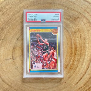 May include: A 1988 Fleer Larry Bird All-Star basketball card, graded and encased in a protective holder. The card features a vibrant image of Larry Bird in action, wearing his Celtics uniform. The card's details include the player's name, the year, and the All-Star designation.