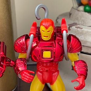 Może przedstawiać: Czerwono-ż&oacute;łta figurka Iron Mana ze srebrnym hakiem na g&oacute;rze. Figurka ma czerwone metaliczne wykończenie z ż&oacute;łtymi akcentami na ramionach i nogach. Figurka trzyma czerwoną metalową broń.
