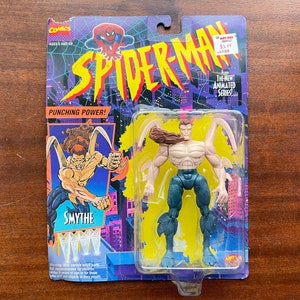 May include: A plastic action figure of the villain Smythe from the Spider-Man animated series. The figure is posed with arms outstretched and has a brown, scaly, textured skin tone. The figure is packaged on a blue, yellow, and purple card with the words "Spider-Man" and "Punching Power!" printed on it.
