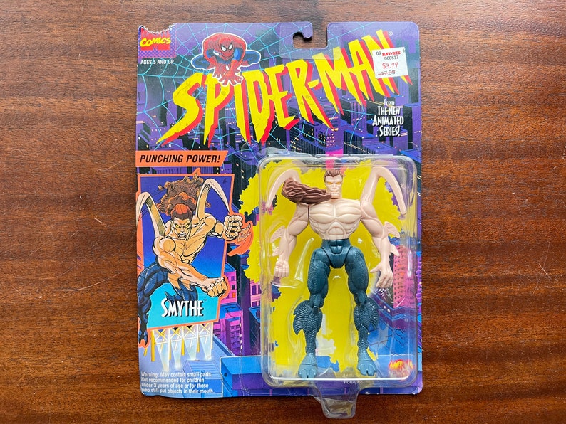 May include: A plastic action figure of the villain Smythe from the Spider-Man animated series. The figure is posed with arms outstretched and has a brown, scaly, textured skin tone. The figure is packaged on a blue, yellow, and purple card with the words "Spider-Man" and "Punching Power!" printed on it.