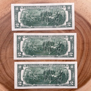 May include: Three two dollar bills featuring a green image of the signing of the Declaration of Independence. The bills are arranged vertically on a wooden surface.