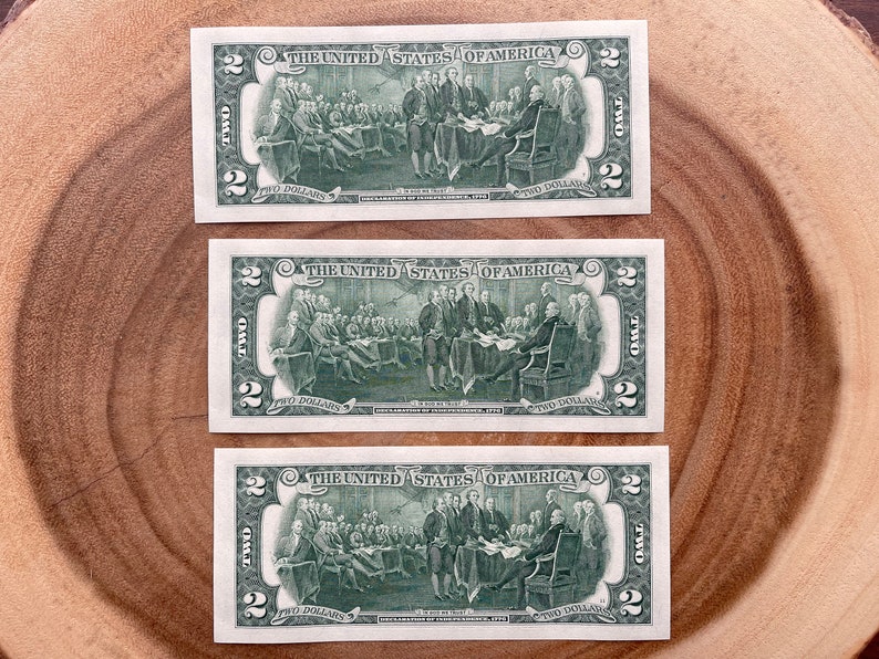 May include: Three two dollar bills featuring a green image of the signing of the Declaration of Independence. The bills are arranged vertically on a wooden surface.