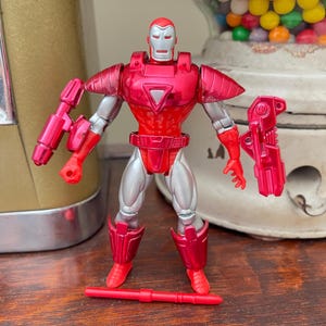 Pode incluir: Figura de a&ccedil;&atilde;o vermelha e prateada de um super-her&oacute;i, com capacete e peitoral vermelhos. A figura tem articula&ccedil;&otilde;es e vem com duas armas destac&aacute;veis. A figura est&aacute; sobre uma superf&iacute;cie de madeira.