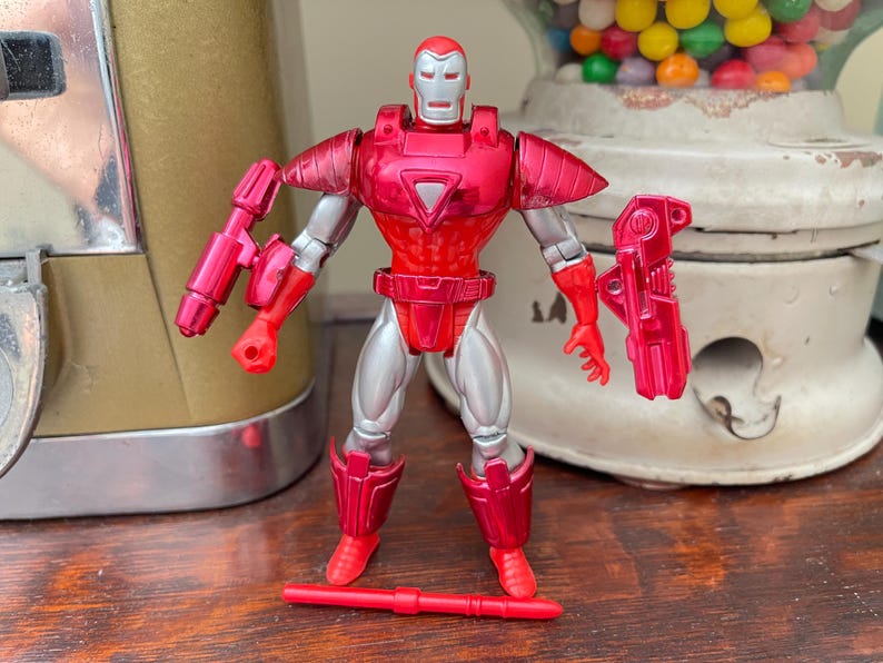 Pode incluir: Figura de a&ccedil;&atilde;o vermelha e prateada de um super-her&oacute;i, com capacete e peitoral vermelhos. A figura tem articula&ccedil;&otilde;es e vem com duas armas destac&aacute;veis. A figura est&aacute; sobre uma superf&iacute;cie de madeira.