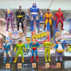 May include: A collection of vintage action figures, including Batman, Superman, The Joker, and Wolverine, displayed on a shelf with baseball card packaging. The figures are arranged in a row, with the packaging behind them. The packaging features the Topps Baseball logo and the words "The Real One!"