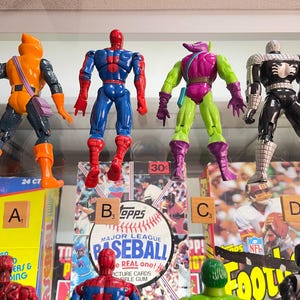 May include: Four action figures of comic book characters are displayed. The figures include a character in orange and black, a blue and red Spider-Man, a green and purple Goblin, and a black and white Spider-Man. The figures are in front of baseball and football cards.