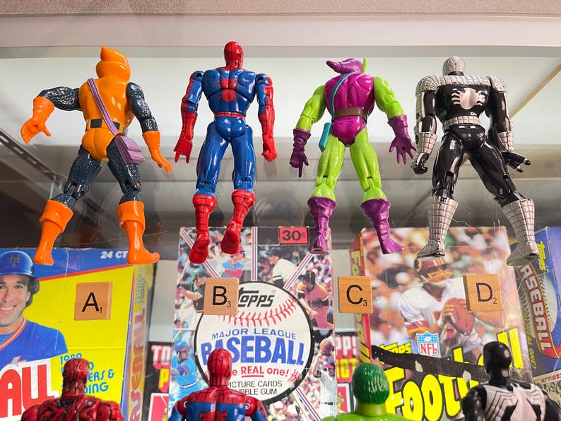 May include: Four action figures of comic book characters are displayed. The figures include a character in orange and black, a blue and red Spider-Man, a green and purple Goblin, and a black and white Spider-Man. The figures are in front of baseball and football cards.