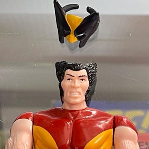 May include: A Wolverine action figure with a removable mask. The figure has a red and yellow torso, tan arms, and black hair. The mask is black and yellow. The figure is a vintage collectible.