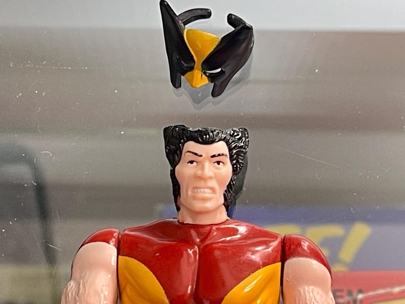 May include: A Wolverine action figure with a removable mask. The figure has a red and yellow torso, tan arms, and black hair. The mask is black and yellow. The figure is a vintage collectible.