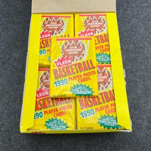 May include: A box of 1990 Fleer Basketball player photo cards. The yellow packs feature a basketball hoop graphic and the text "Fifth Anniversary Edition." The box is yellow and the packs are arranged inside.