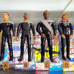 May include: Four Star Trek action figures in black uniforms with silver and gold accents. The figures stand in front of baseball and football card boxes. Scrabble tiles spell out A, B, C, and D. The figures are approximately 6 inches tall.
