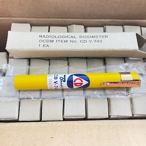 May include: A yellow radiological dosimeter with a gold clip, packaged in a box. The dosimeter has a blue, white, and red logo and the text "CD V-742". The box contains multiple small, square cardboard containers.