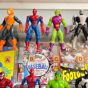 May include: Four action figures of Marvel comic characters, including Spider-Man, Hobgoblin, Green Goblin, and Spider-Man in a black and white suit. The figures are displayed in front of baseball and football card boxes.