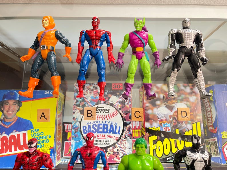 May include: Four action figures of Marvel comic characters, including Spider-Man, Hobgoblin, Green Goblin, and Spider-Man in a black and white suit. The figures are displayed in front of baseball and football card boxes.