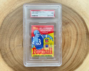 May include: A graded 1985 Topps Football wax pack. The pack features a colorful design with a football player illustration and the word "Football." The pack is encased in a clear protective holder, with a label indicating the grade and serial number.
