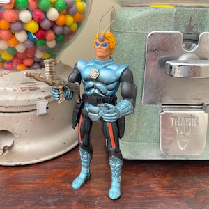 May include: A blue and black action figure with orange hair and a silver weapon. The figure is standing in front of a vintage gumball machine and a green vending machine with a "THANK YOU" sign.