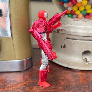 Pode incluir: Figura de a&ccedil;&atilde;o vermelha e prateada de um super-her&oacute;i, com acabamento met&aacute;lico. A figura est&aacute; posando com uma arma, perto de uma m&aacute;quina de chicletes vintage e um recipiente de vidro cheio de doces.