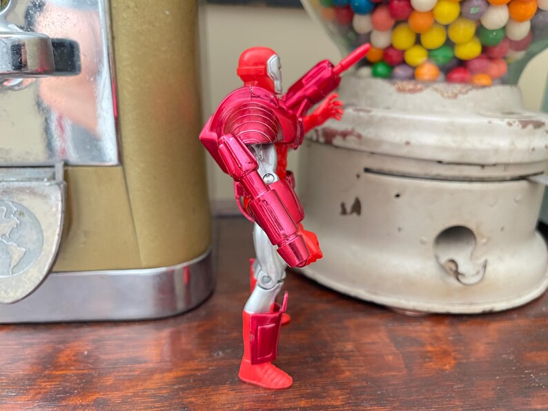Pode incluir: Figura de a&ccedil;&atilde;o vermelha e prateada de um super-her&oacute;i, com acabamento met&aacute;lico. A figura est&aacute; posando com uma arma, perto de uma m&aacute;quina de chicletes vintage e um recipiente de vidro cheio de doces.