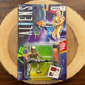 May include: Action figure of a space marine with a gun, holding a comic book. The figure is wearing a green and brown uniform and a helmet. The background is a blue and purple alien landscape. The packaging says "Aliens" and "Space Marine Drake".