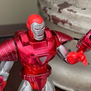 Pode incluir: Figura de a&ccedil;&atilde;o vermelha e prateada de um super-her&oacute;i com capacete e armadura. A figura tem um acabamento met&aacute;lico e est&aacute; posada com um bra&ccedil;o estendido, mostrando uma arma. O design da figura &eacute; detalhado e estilizado.