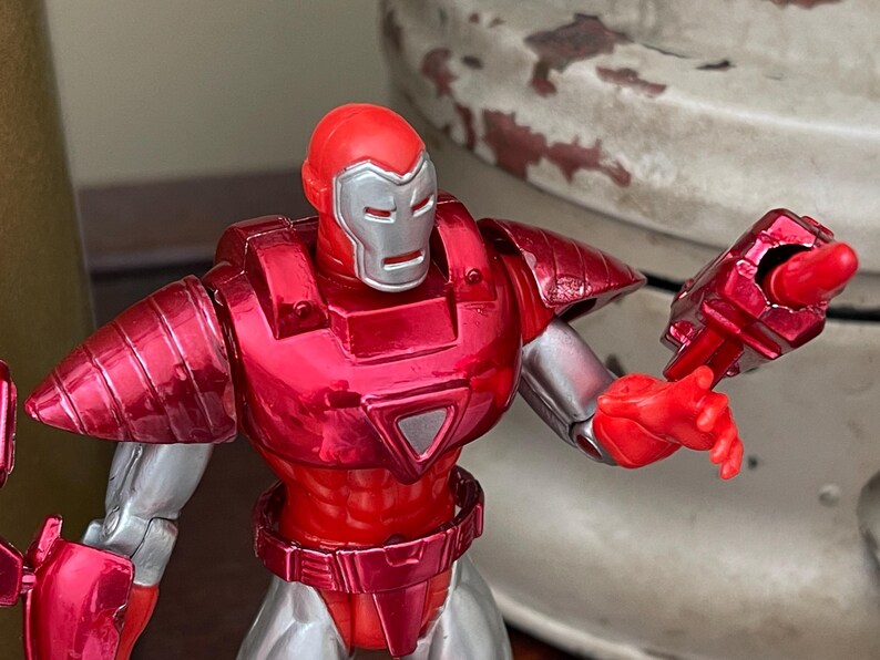 Pode incluir: Figura de a&ccedil;&atilde;o vermelha e prateada de um super-her&oacute;i com capacete e armadura. A figura tem um acabamento met&aacute;lico e est&aacute; posada com um bra&ccedil;o estendido, mostrando uma arma. O design da figura &eacute; detalhado e estilizado.