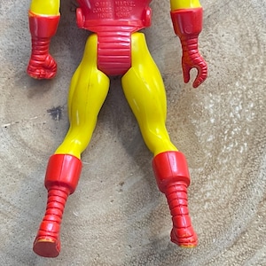May include: A vintage Iron Man action figure, featuring a red and yellow color scheme. The figure's legs are yellow, with red boots and a red torso. The arms are red. The figure is made of plastic and shows signs of wear.