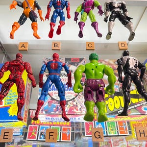 May include: A collection of eight action figures of comic book characters, including Spider-Man, Hulk, and Venom. The figures are in various poses and colors, displayed in a glass case. Scrabble tiles with letters are placed below the figures.