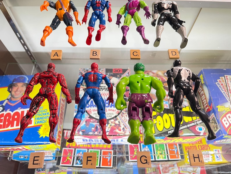 May include: A collection of eight action figures of comic book characters, including Spider-Man, Hulk, and Venom. The figures are in various poses and colors, displayed in a glass case. Scrabble tiles with letters are placed below the figures.