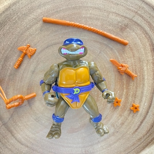 May include: A Teenage Mutant Ninja Turtle action figure with a blue mask, orange belt, and various orange weapons. The figure is brown and yellow, with blue accents. Accessories include a bo staff, sai, and throwing stars.