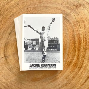 May include: A stack of baseball cards featuring Jackie Robinson in a black and white image. The card shows Robinson in a baseball uniform with his arms raised. The text "JACKIE ROBINSON INFIELDER-BROOKLYN DODGERS" is printed at the bottom of the card.
