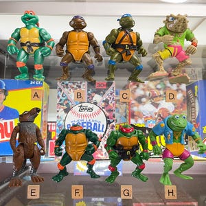May include: Collection of vintage action figures, including Teenage Mutant Ninja Turtles and a Splinter figure. The figures are displayed in a glass case, with baseball card boxes in the background. The figures are in various colors.