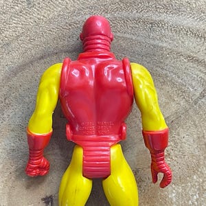 May include: A vintage Iron Man action figure, viewed from the back. The figure is primarily red and yellow, with a red helmet, back, and accents on the arms and legs. The arms and legs are yellow. The figure is made of plastic.