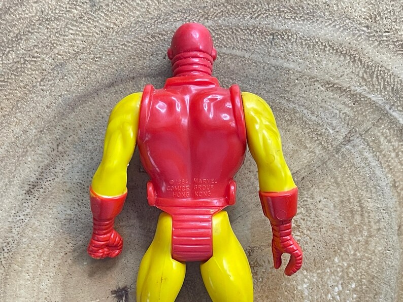 May include: A vintage Iron Man action figure, viewed from the back. The figure is primarily red and yellow, with a red helmet, back, and accents on the arms and legs. The arms and legs are yellow. The figure is made of plastic.